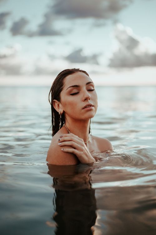 Free Portrait of a relaxed woman with eyes closed standing in calm ocean water at sunset. Stock Photo