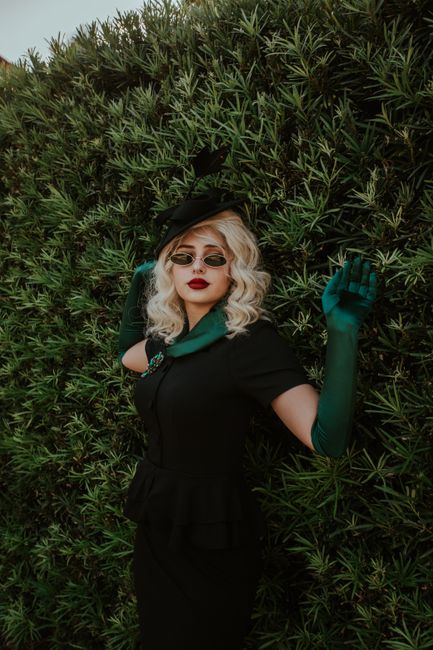 Stylish woman in vintage attire with sunglasses posing by a lush hedge.