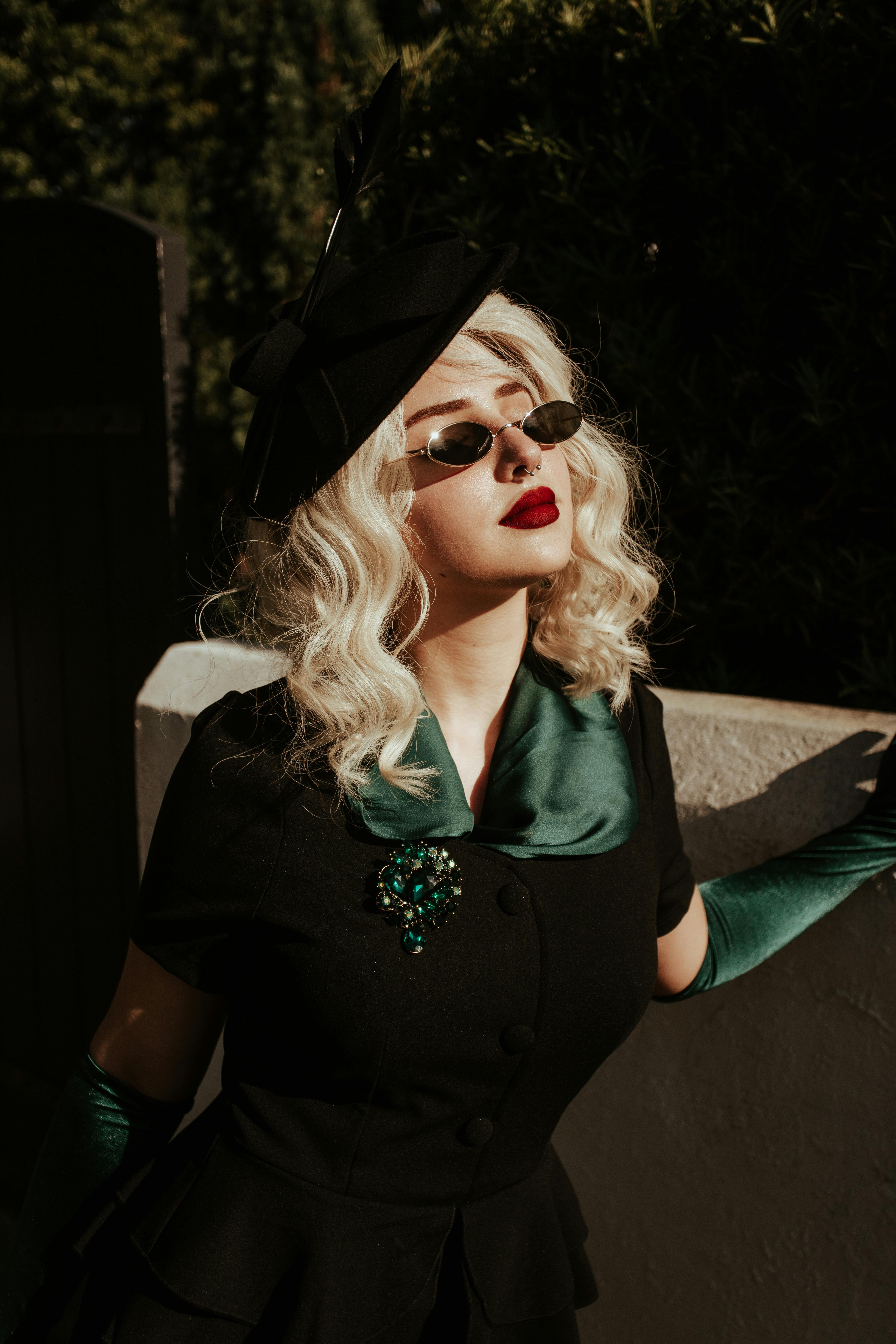 Elegant woman in vintage attire poses outdoors with striking red lipstick and dark sunglasses.