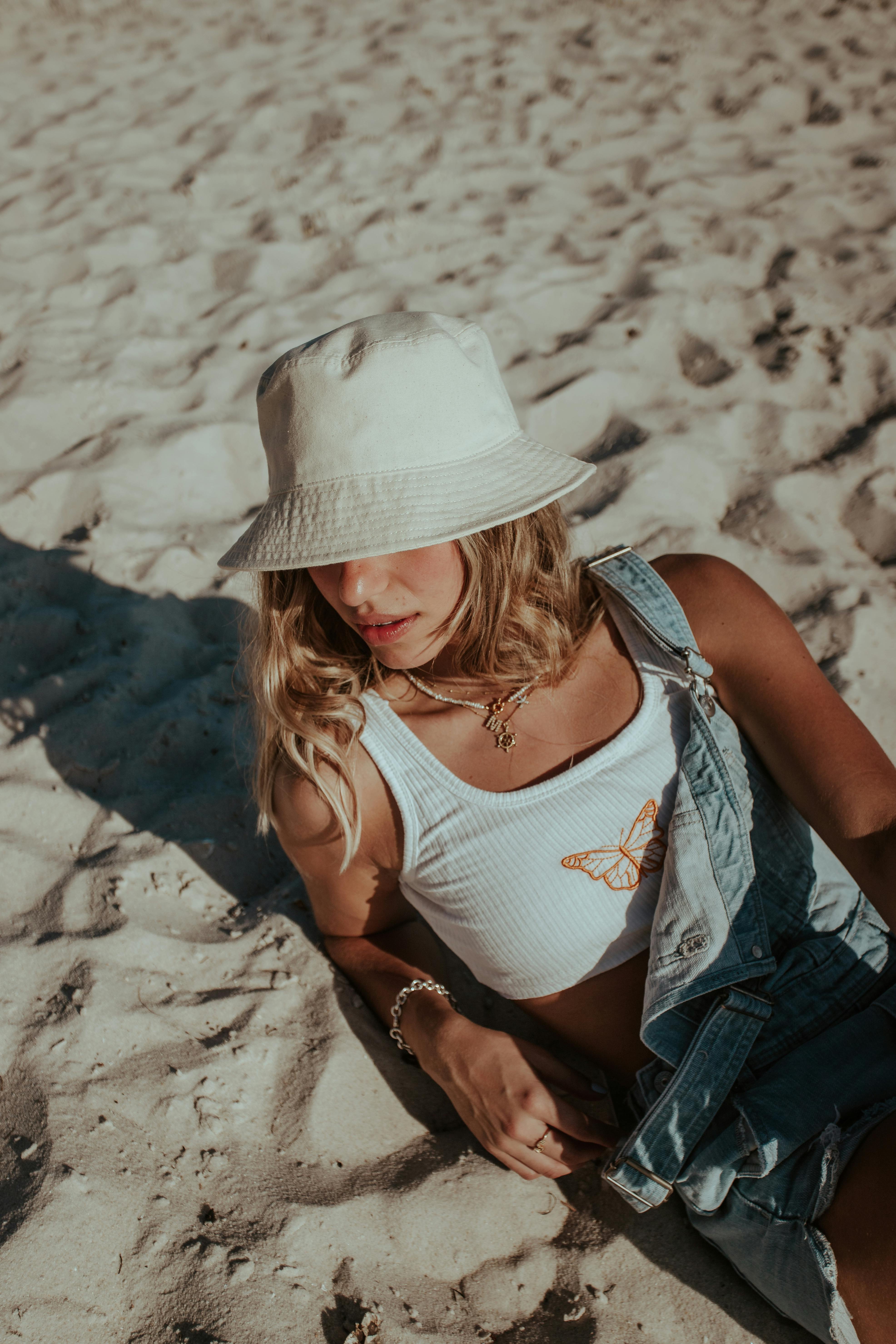 Woman in denim overalls and sun hat relaxing on sandy beach in summer sun.