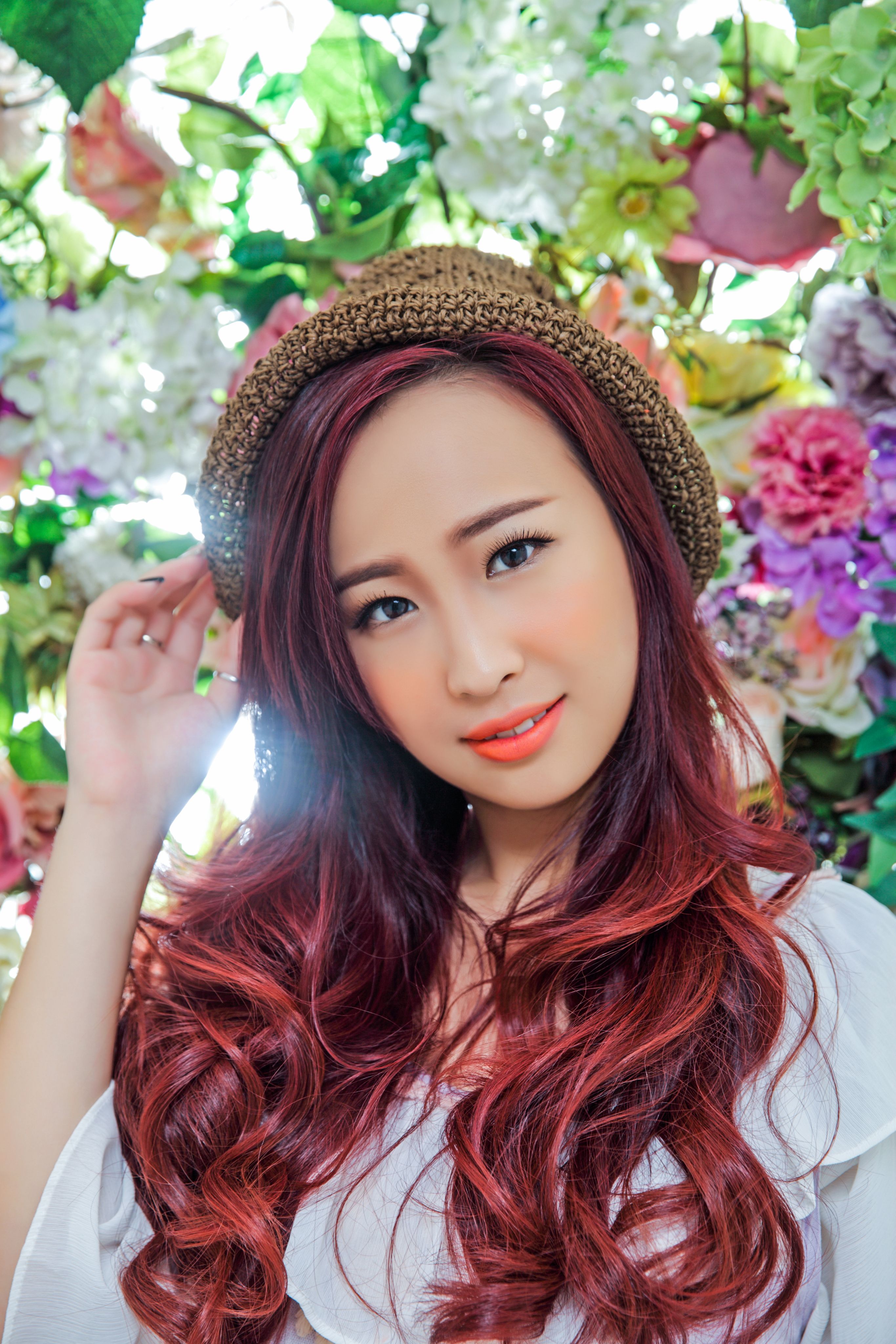 Asian woman with red hair posing in front of colorful floral wall, wearing a hat.