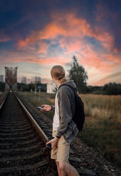 A man in casual attire stands on railway tracks at sunset, holding a camera and a smartphone.