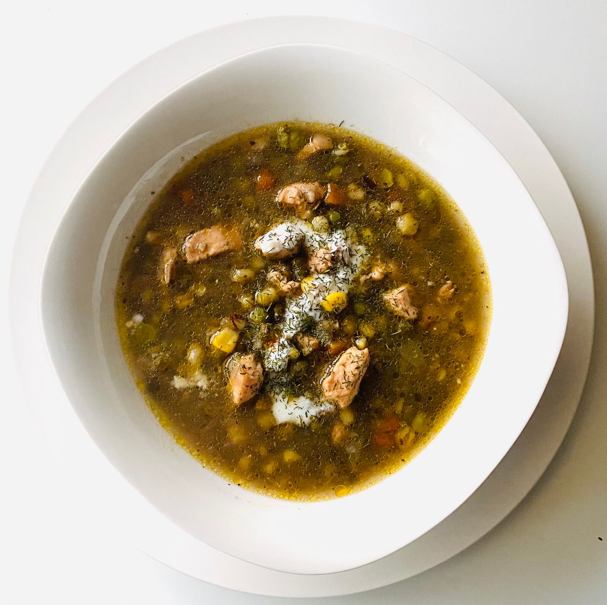 Overhead bowl of chicken soup with peas, corn, carrot, sour cream and chopped dill