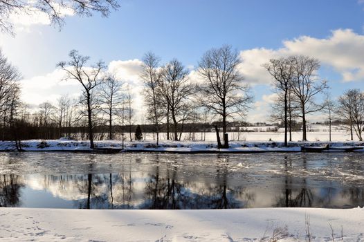 A serene snowy winter scene with a frozen river and bare trees under a blue sky.