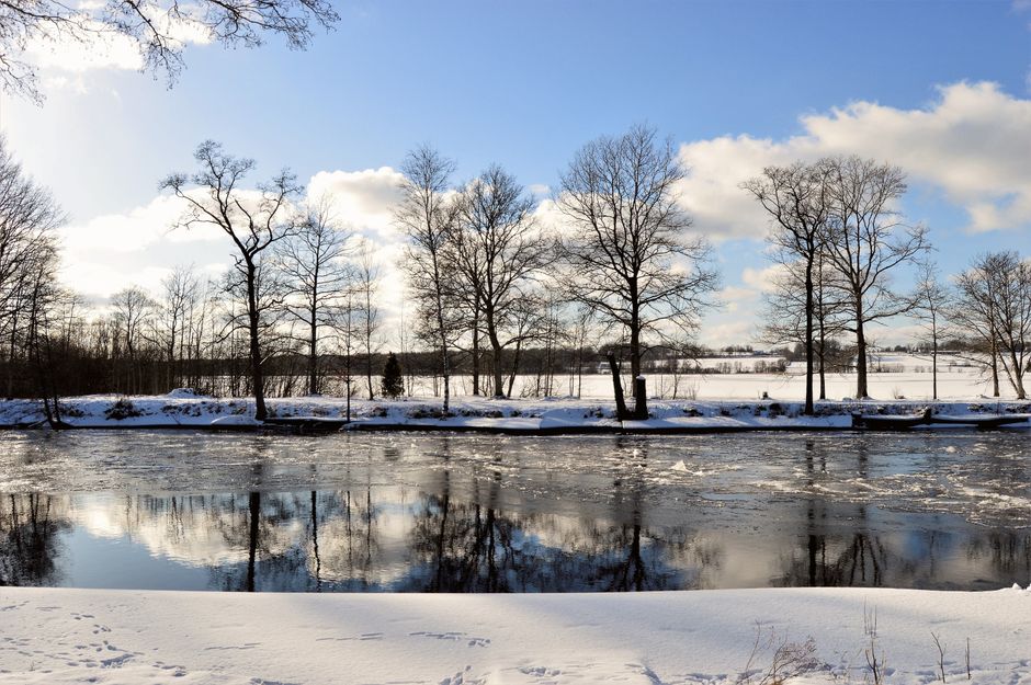 A serene snowy winter scene with a frozen river and bare trees under a blue sky.