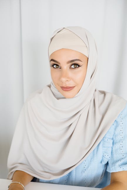 A beautiful woman in a hijab smiles warmly while seated indoors against a white background.