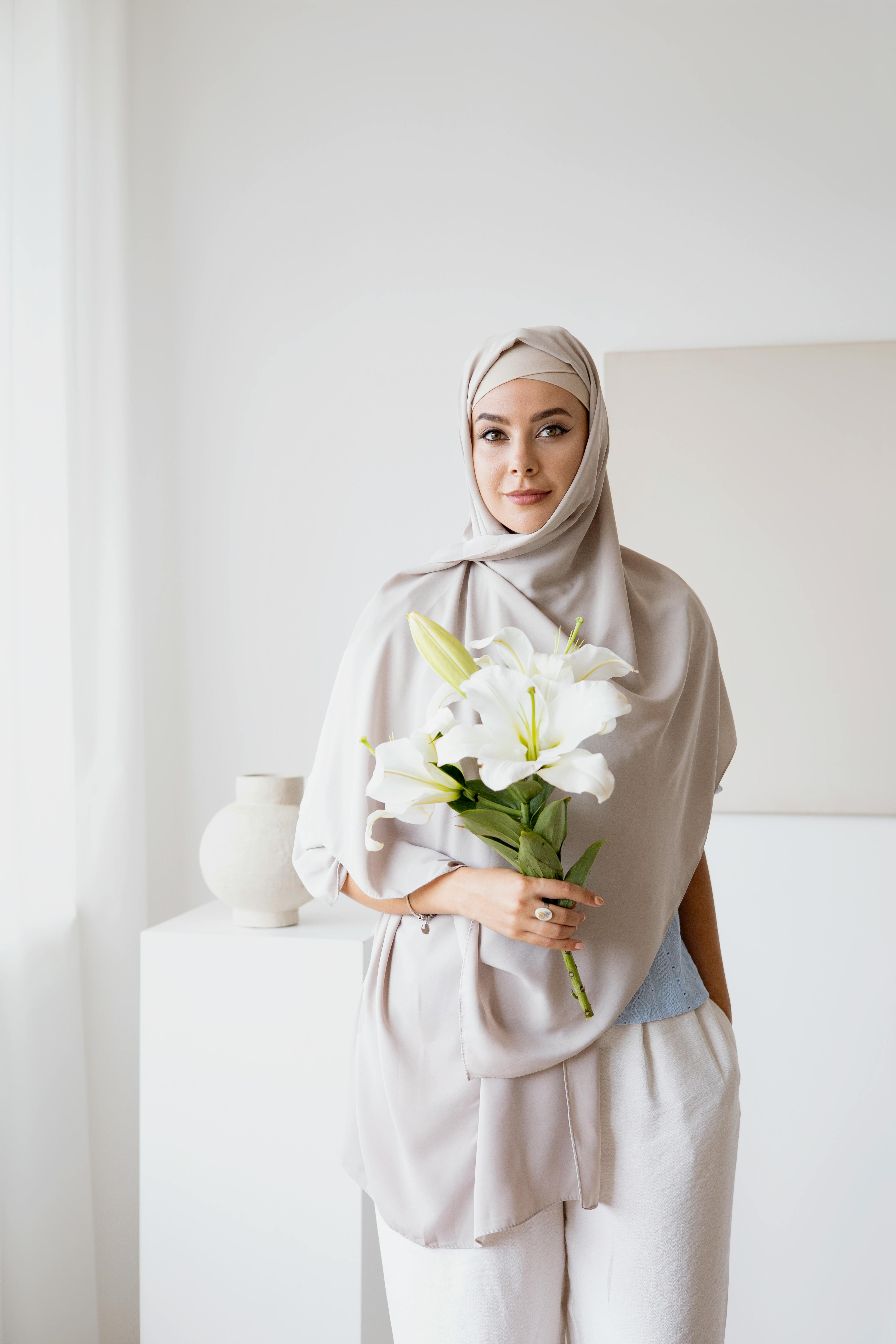 Portrait of a woman in a hijab holding white lilies in a minimalist indoor setting.