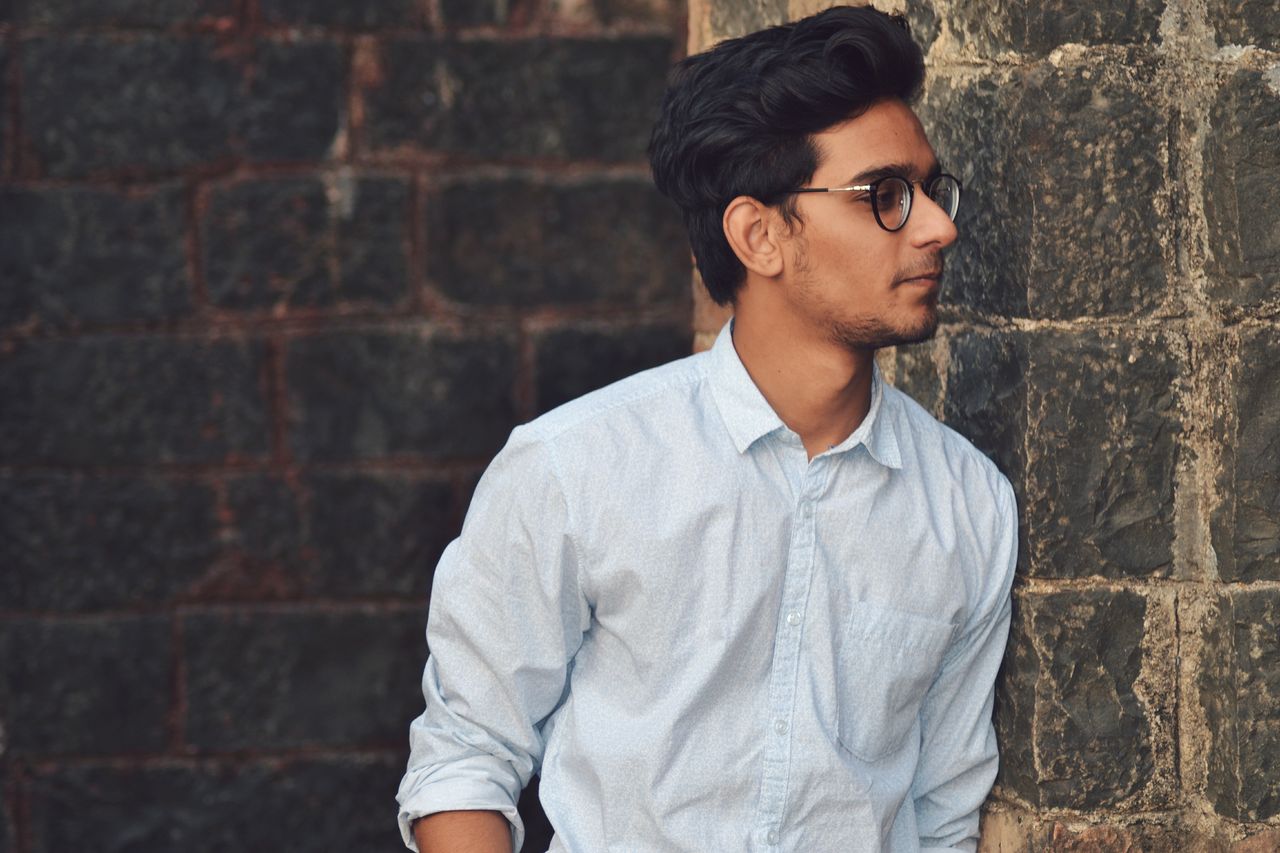 a stylish young man in glasses leans against a brick wall wearing a ca