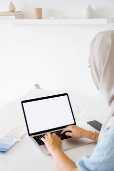 A woman in a hijab working remotely on a laptop at a bright, minimalist indoor setting.