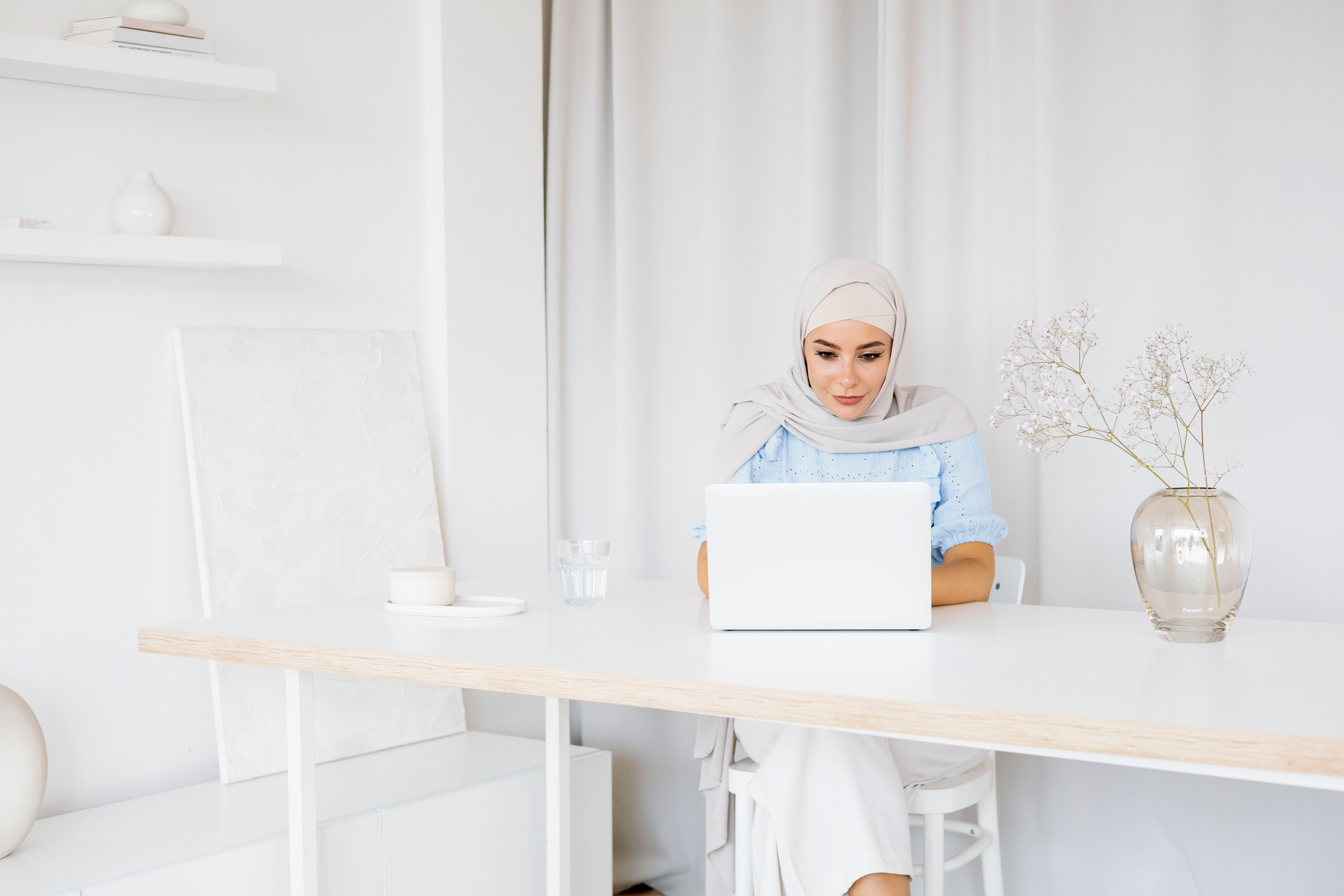 A woman in a hijab sits at a white desk using a laptop, showcasing a calm home workspace.