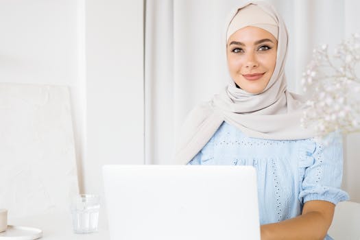 A smiling woman in a hijab using a laptop indoors with a bright and calm setting.