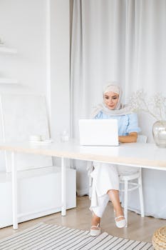 Woman in hijab engaged in remote work at home using a laptop. Bright and modern workspace indoors.