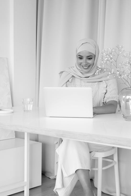 Free Grayscale Photo of a Woman with Hijab Using a Laptop while Sitting Stock Photo