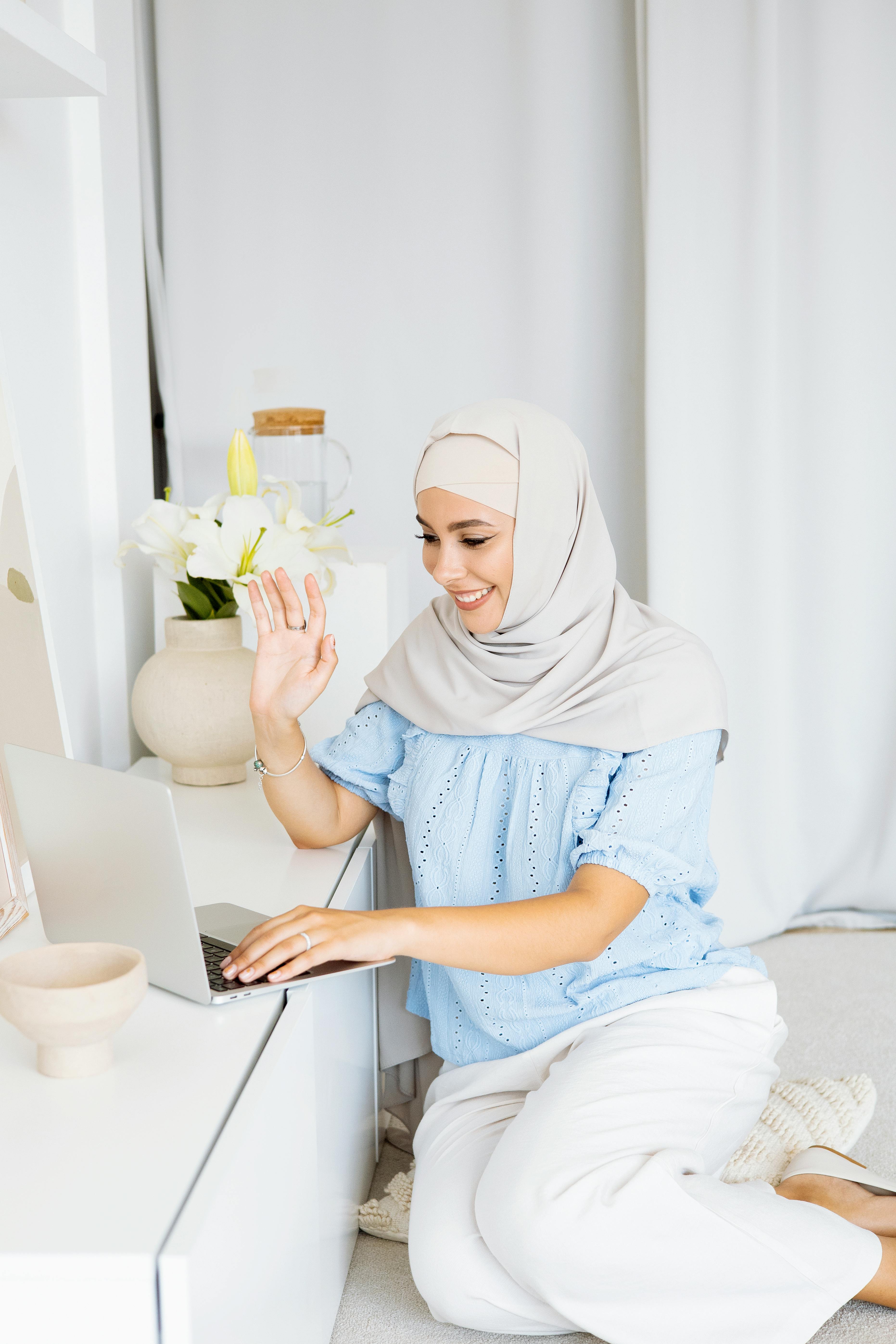 Smiling woman in hijab sitting on floor, engaged in remote work using a laptop.