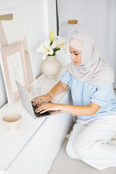 Young Muslim woman in hijab typing on her laptop indoors, surrounded by modern decor.