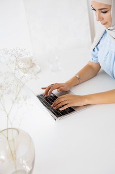 Muslim woman in hijab typing on a laptop in a bright, minimalist setting.