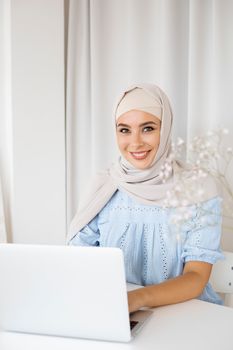 Confident woman in hijab sitting at a wooden table using a laptop for remote work.