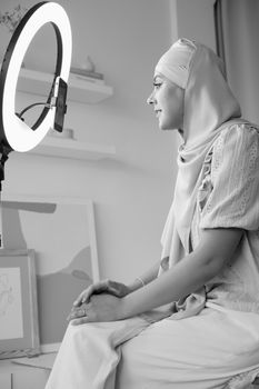 Muslim woman wearing hijab sitting indoors vlogging with ring light and smartphone.