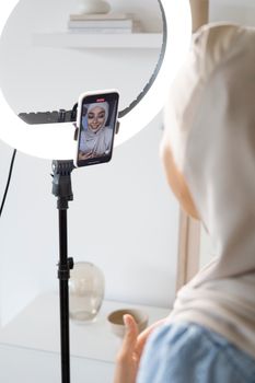 Muslim woman using smartphone and ring light for vlogging indoors.