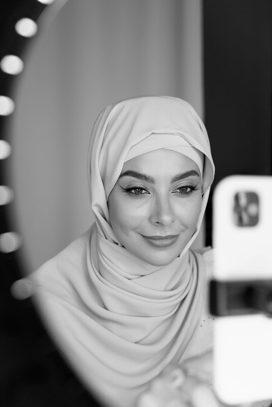 Black and white portrait of a woman in hijab smiling at her phone indoors.