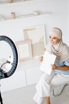 Woman in hijab using ring light indoors for creating lifestyle vlog content.