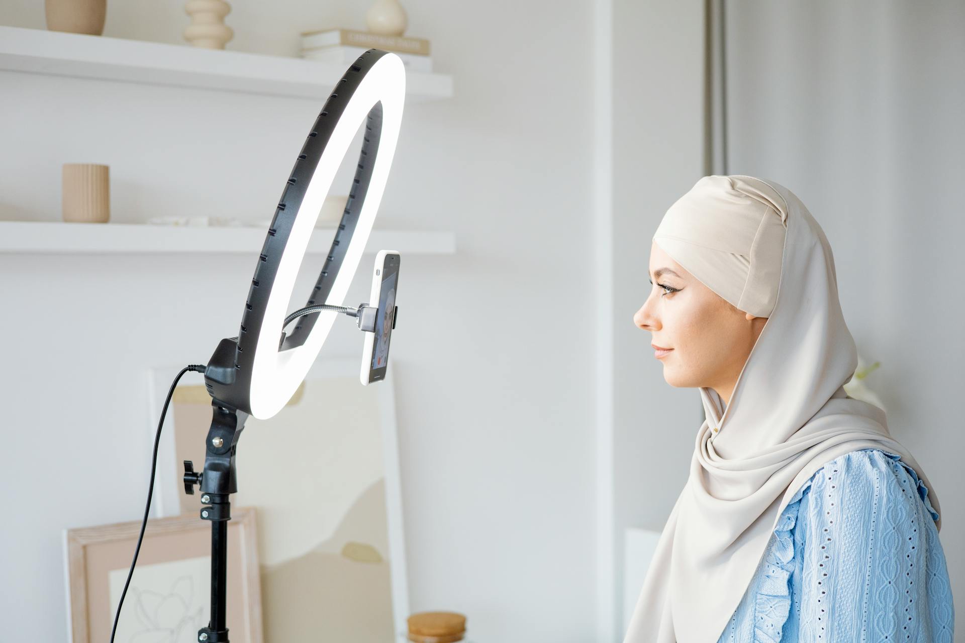 A woman in hijab uses ring light for video content creation indoors.