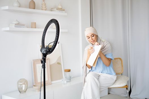 Woman wearing a hijab holding a clipboard while recording an online teaching session with a ring light.