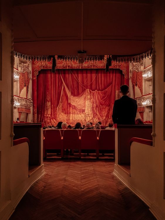 View from an aisle seat in an elegant theater with red velvet curtains and audience members seated