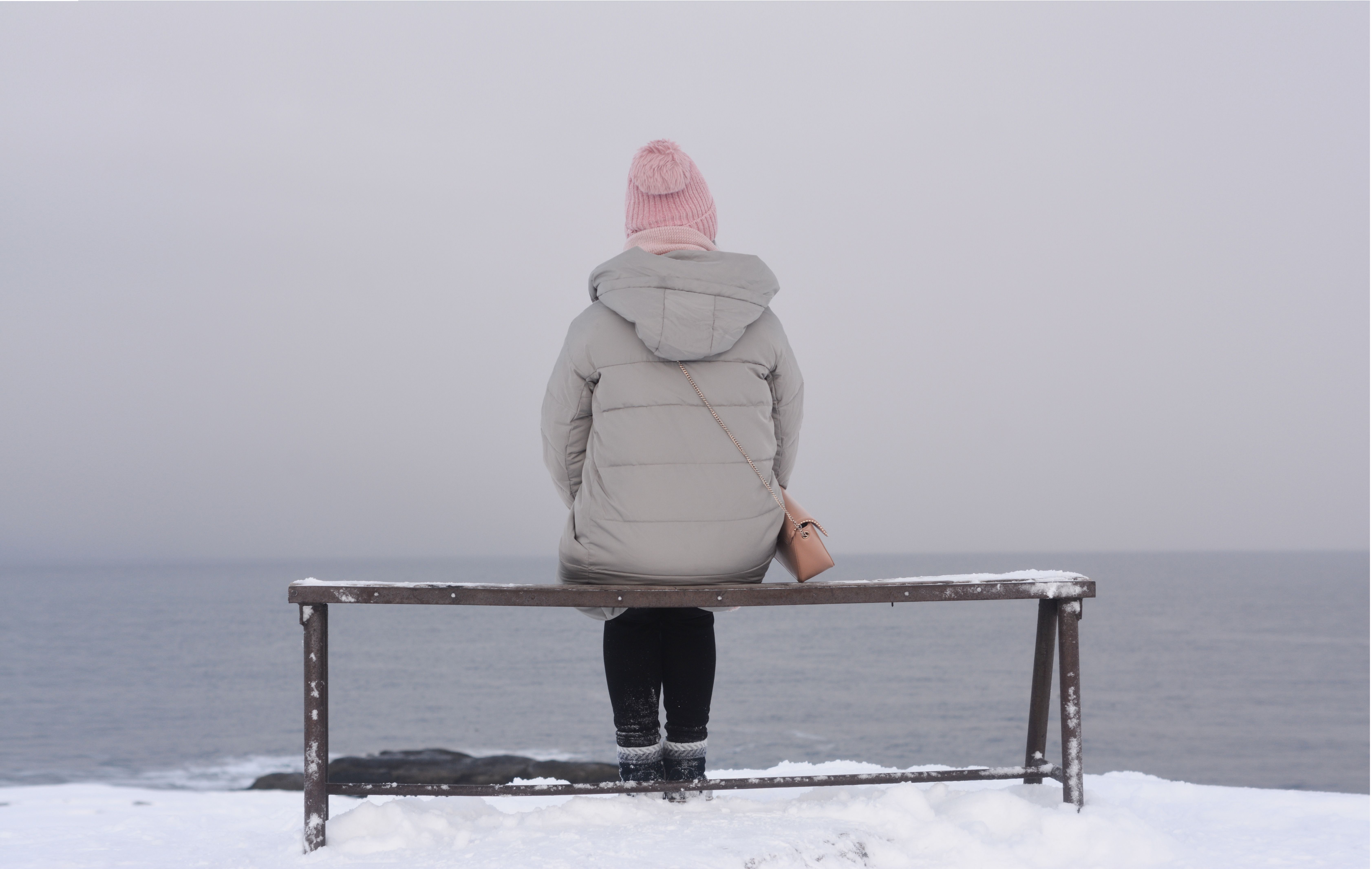 A person sits on a bench facing the snowy sea, wearing a beanie and jacket.