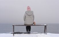 A person sits on a bench facing the snowy sea, wearing a beanie and jacket.