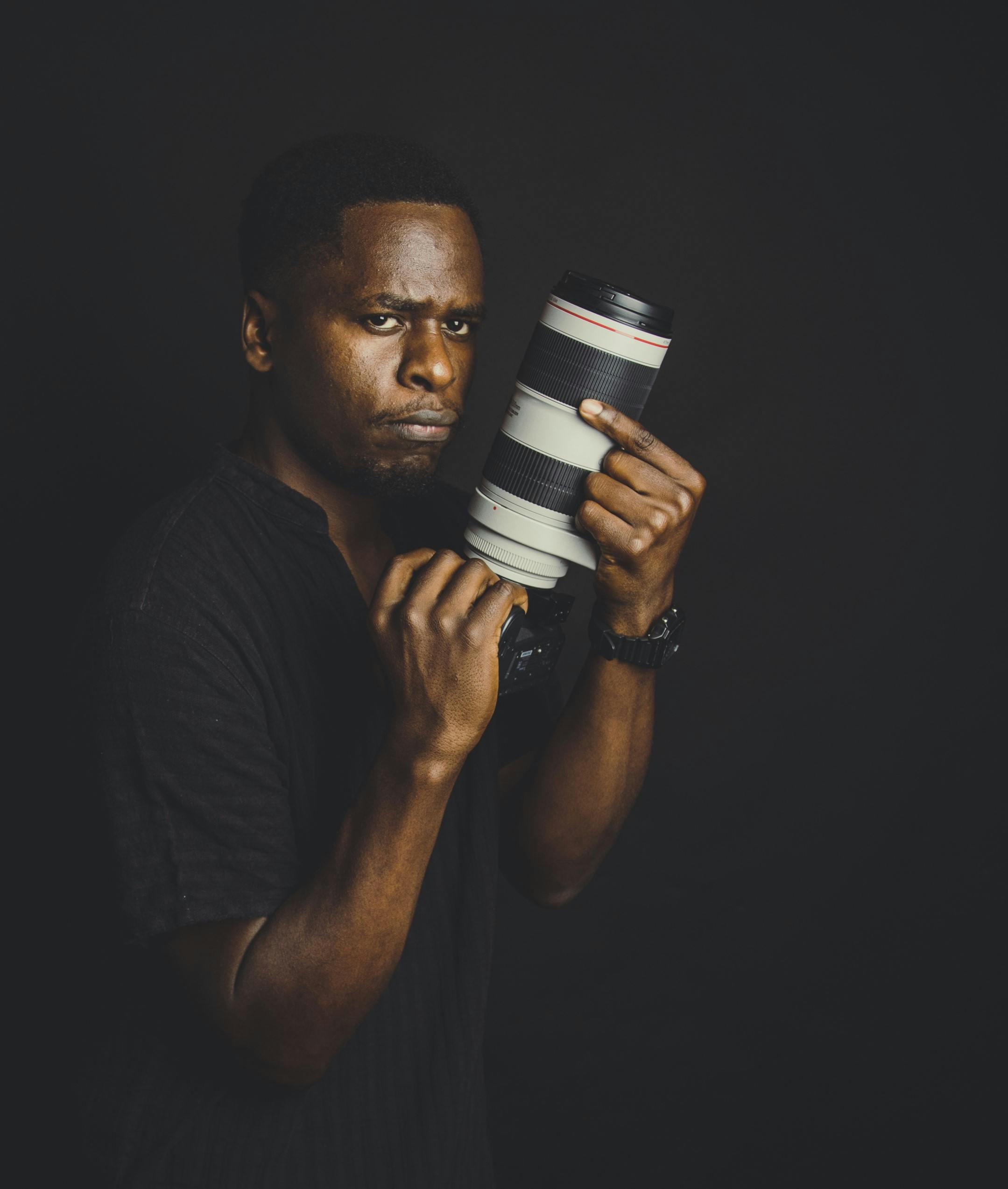 Portrait of a serious photographer holding a DSLR with a large lens, set against a black studio background.