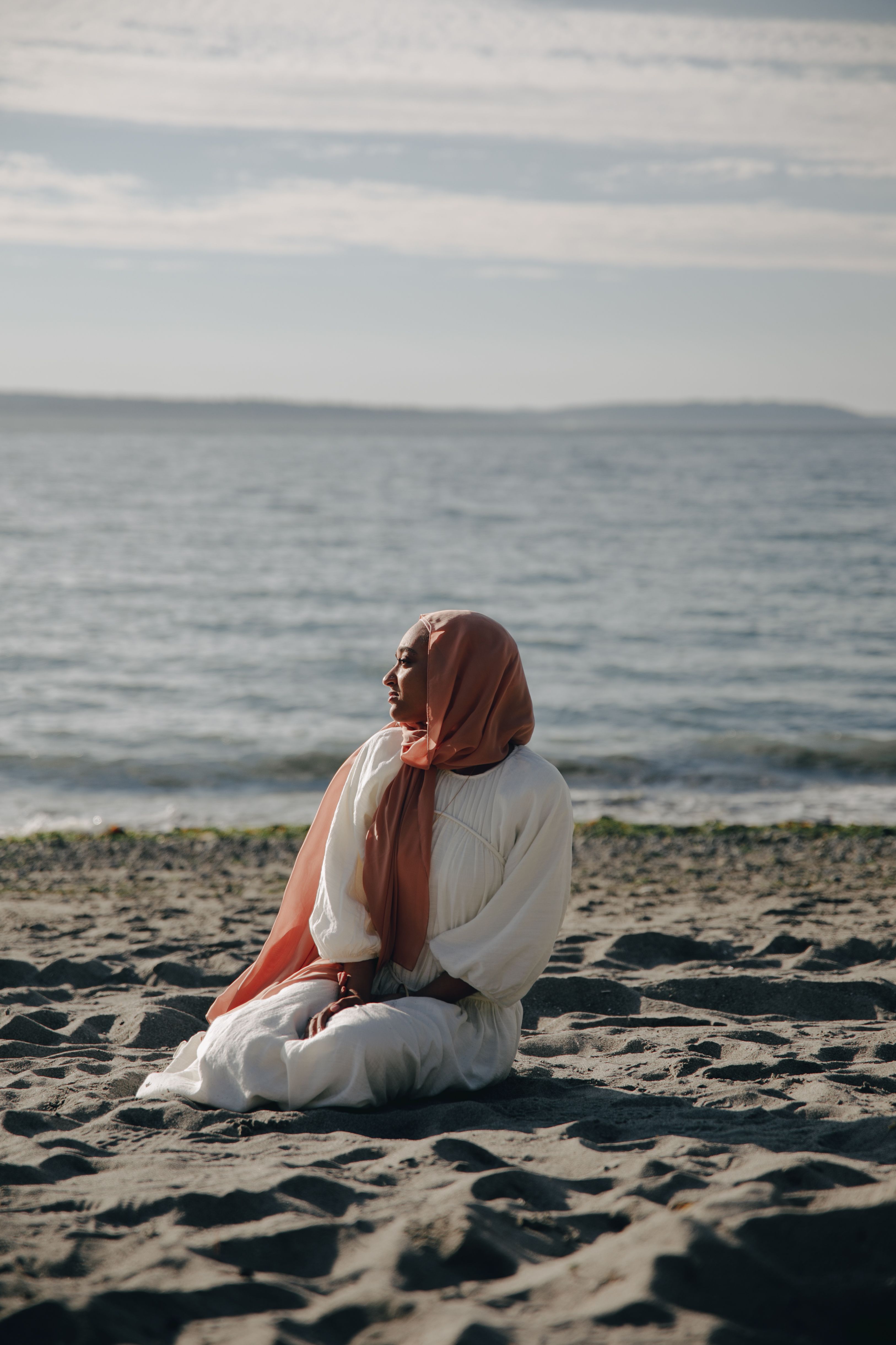 Peaceful scene of a woman in hijab sitting on a sandy beach, reflecting by the sea.