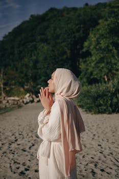 Side view of a woman in a headscarf praying on a beach, serene and peaceful setting.