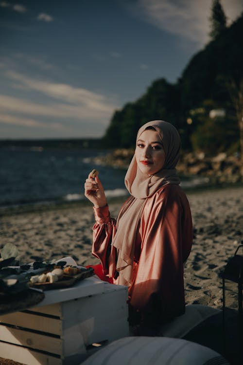A Woman Wearing a Hijab Sitting on Shore Holding a Cookie