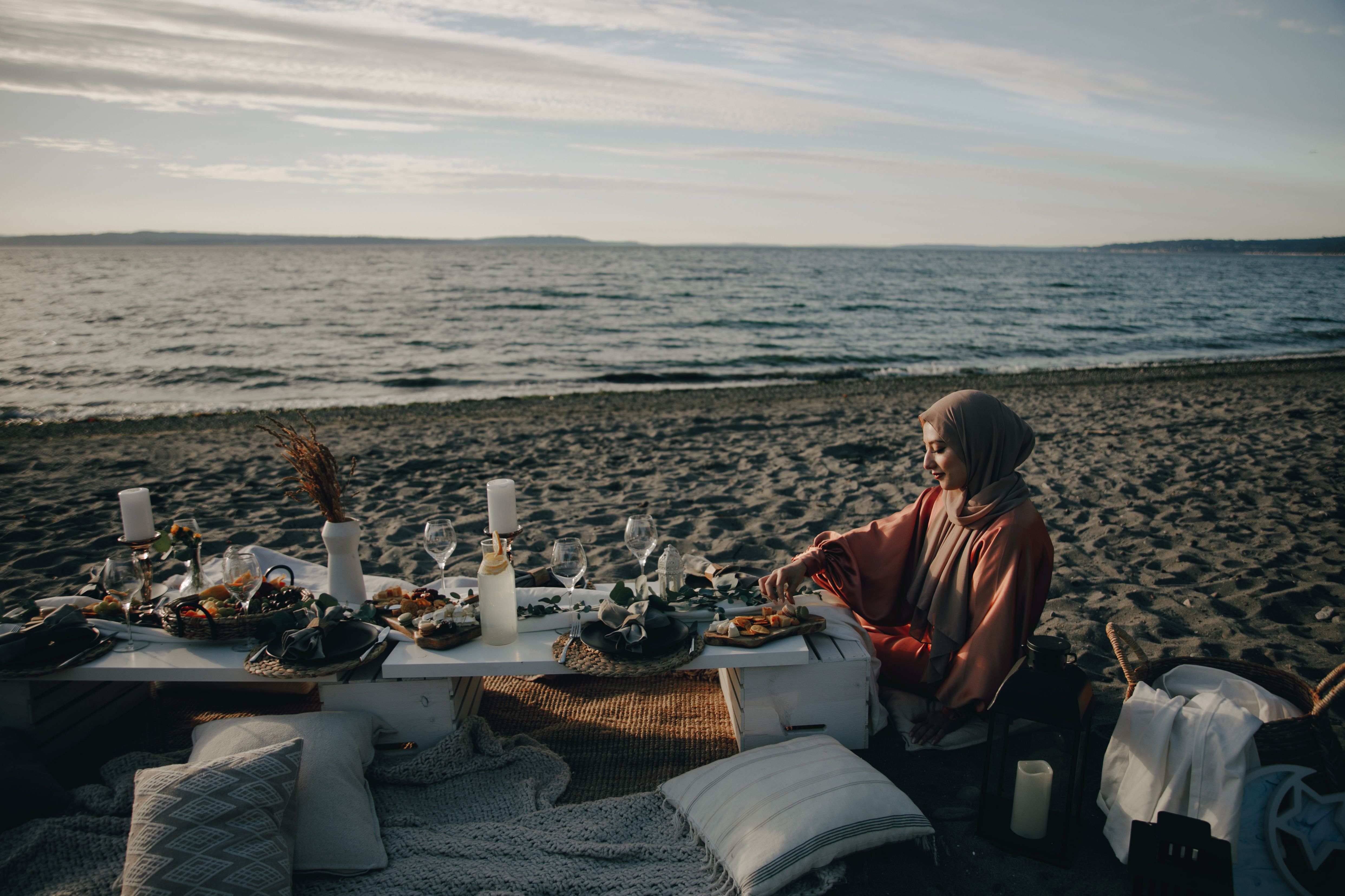 A woman in a hijab enjoys a peaceful beach picnic at sunset, surrounded by food and ocean views.