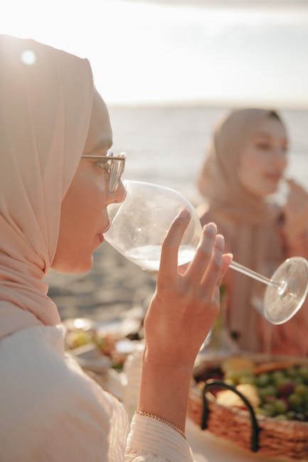 Two Muslim women in hijabs enjoy a peaceful picnic by the beach at sunset, sipping drinks and relaxing.