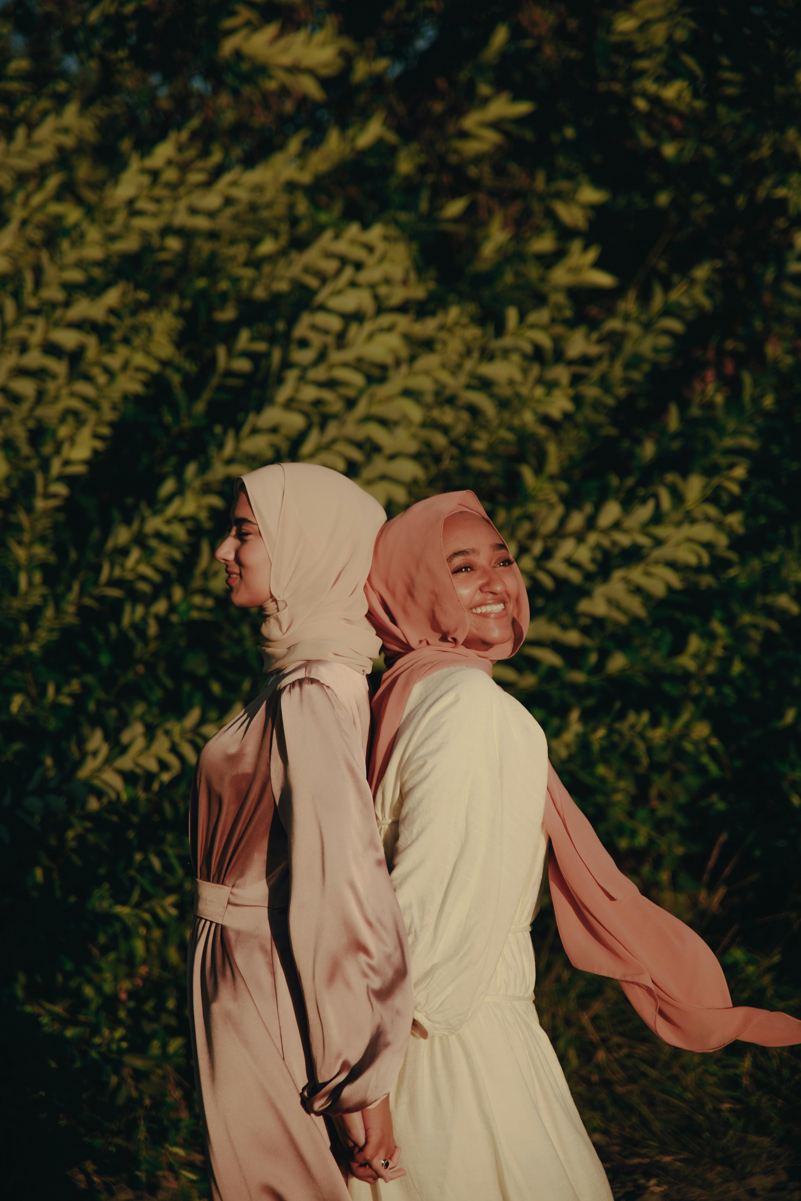 two women in hijab and dresses standing back to back beside tree during sunset