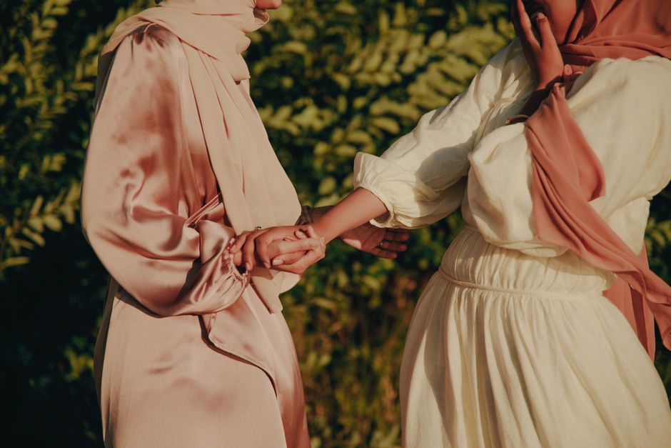 Two women dressed in elegant Muslim fashion holding hands outdoors. Warm tones and soft lighting.
