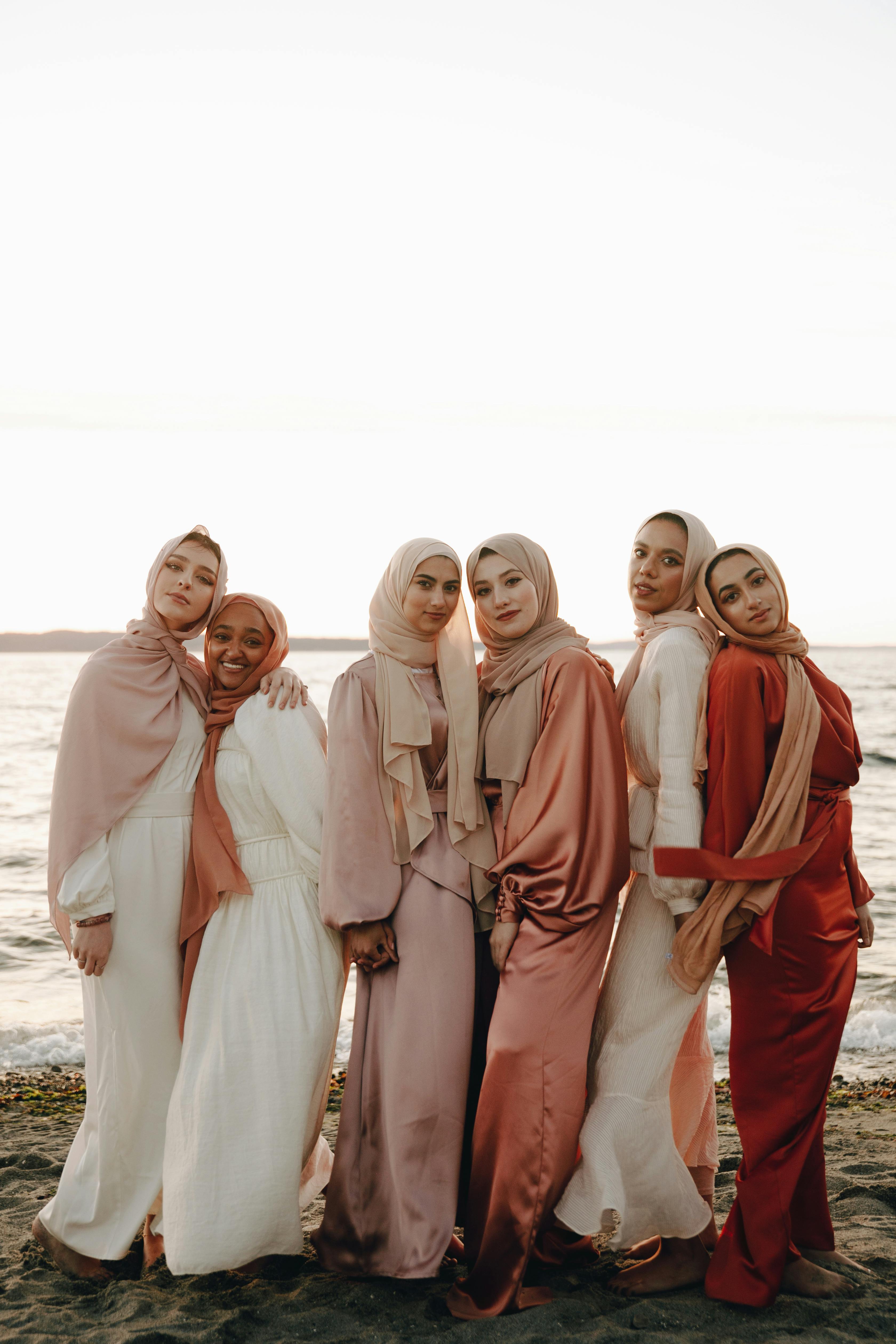 A group of women in elegant dresses and hijabs enjoying a beach sunset.