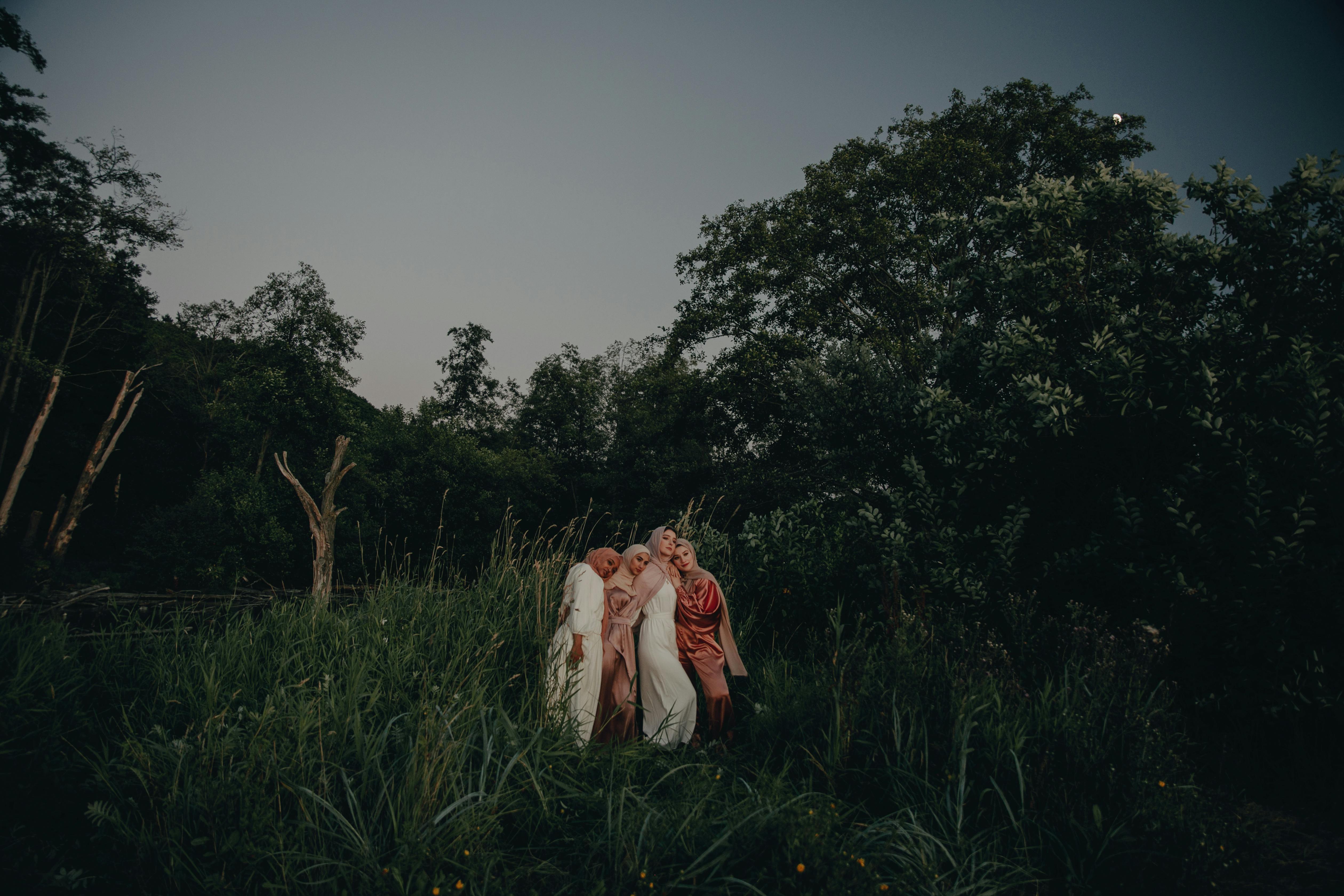 Four women in hijabs embracing each other amidst lush greenery at dusk, creating a serene atmosphere.