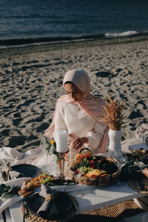 Free A peaceful beach picnic with a woman in hijab, surrounded by fruits and candles, during the day. Stock Photo