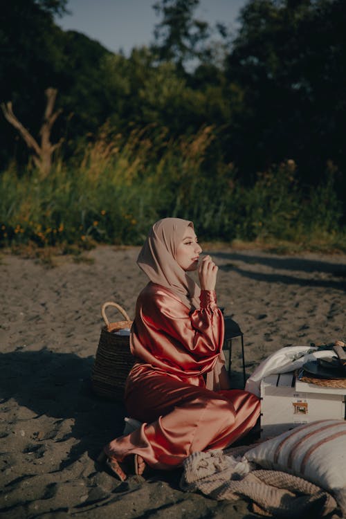 Woman in Pink Satin Abaya and Brown Hijab Sitting on Beach Sand