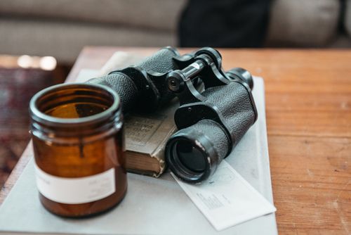 Free A close-up of vintage binoculars resting on a wooden table with a book and amber glass jar. Stock Photo