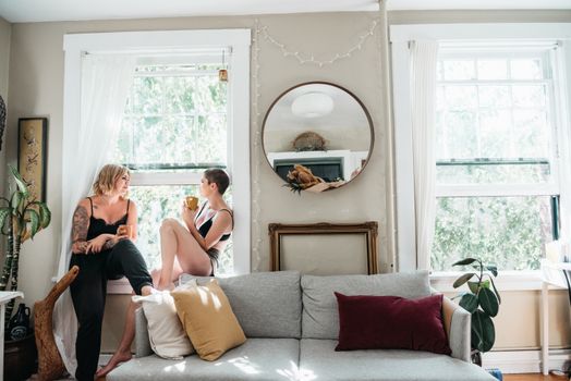 A lesbian couple enjoys a peaceful moment with coffee on a window sill, inside a cozy living room.