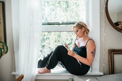 A Woman Sitting on the Window Sill Writing on a Paper