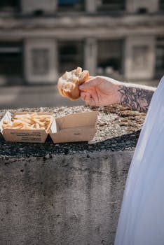 Hand holding a burger with fries in a box on a sunlit urban ledge.