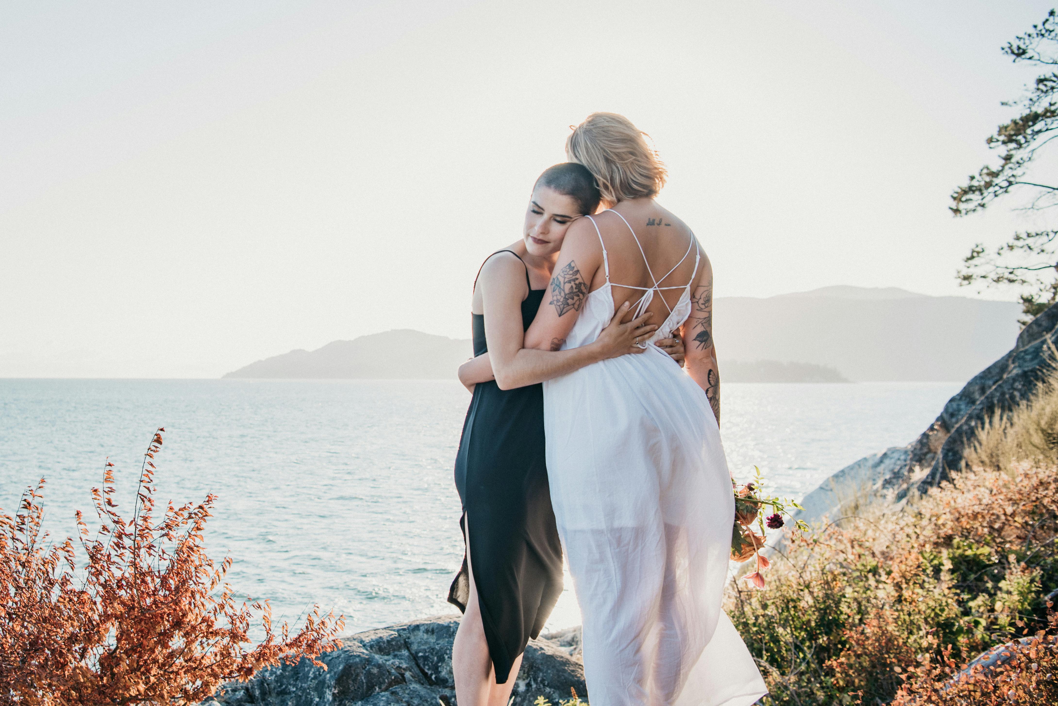 A couple embraces by the ocean, showcasing tattoos and love against a scenic backdrop.