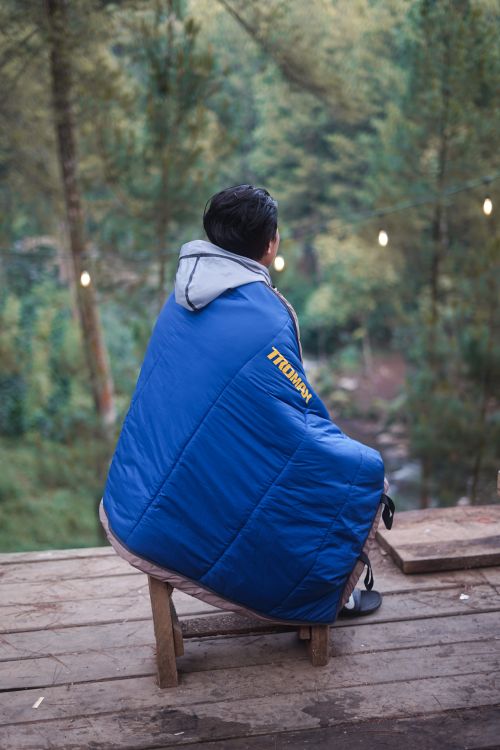 Free A person sits wrapped in a blue blanket on a wooden deck overlooking lush forest scenery. Stock Photo