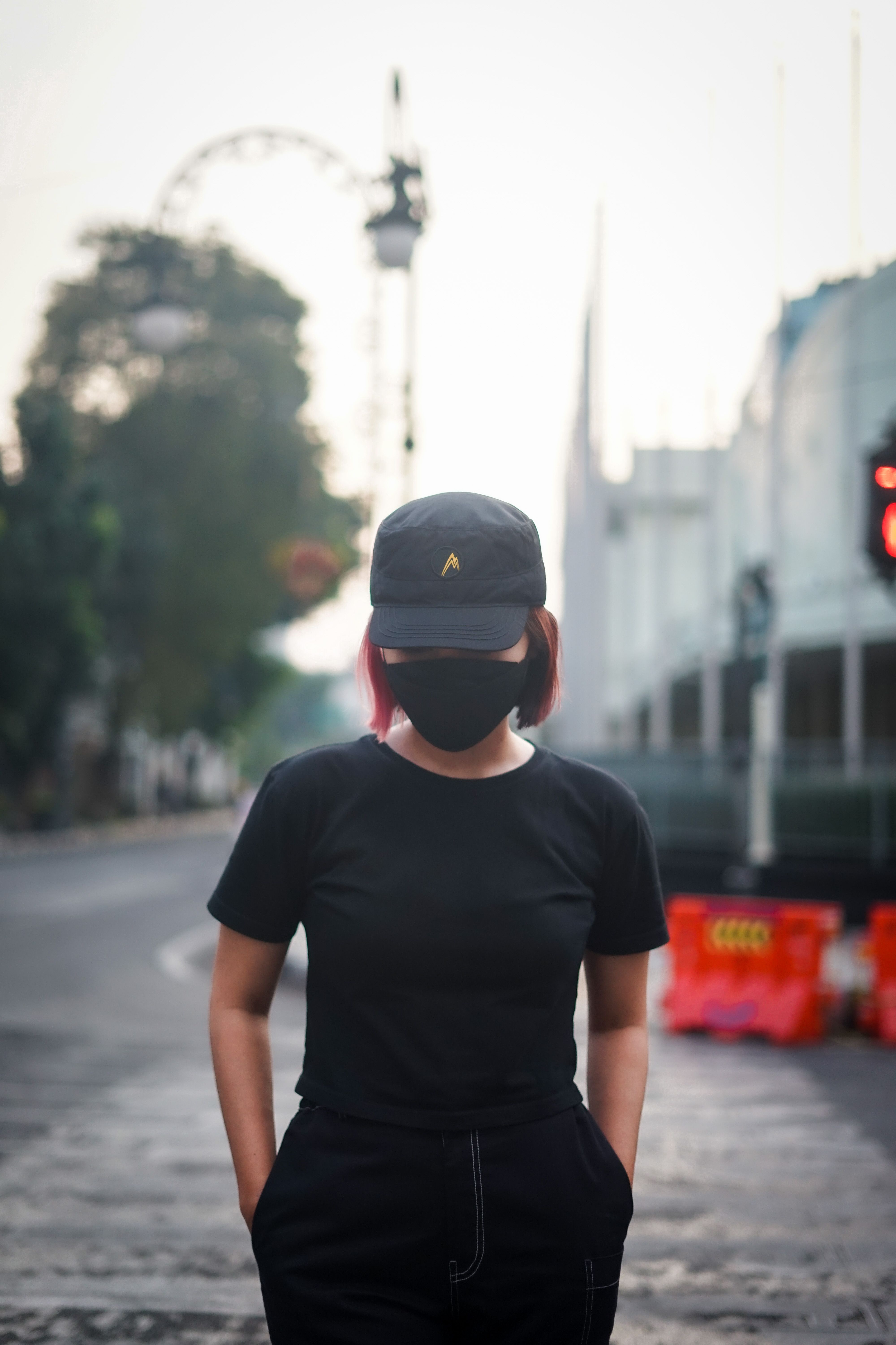 A woman in black clothes with a mask and cap standing on a city street.