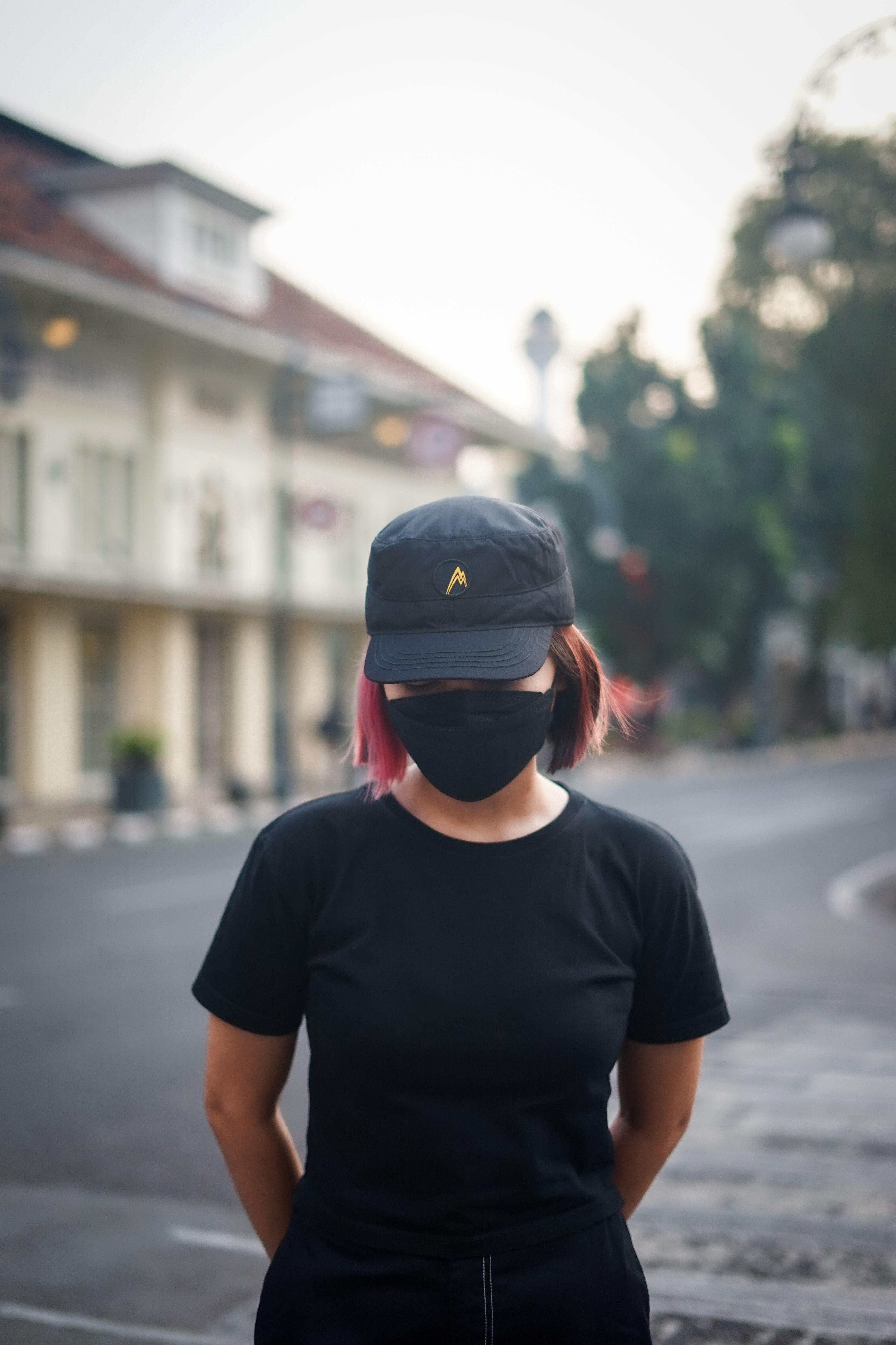 A woman wearing a black cap and mask stands on a city street, creating a modern urban vibe.